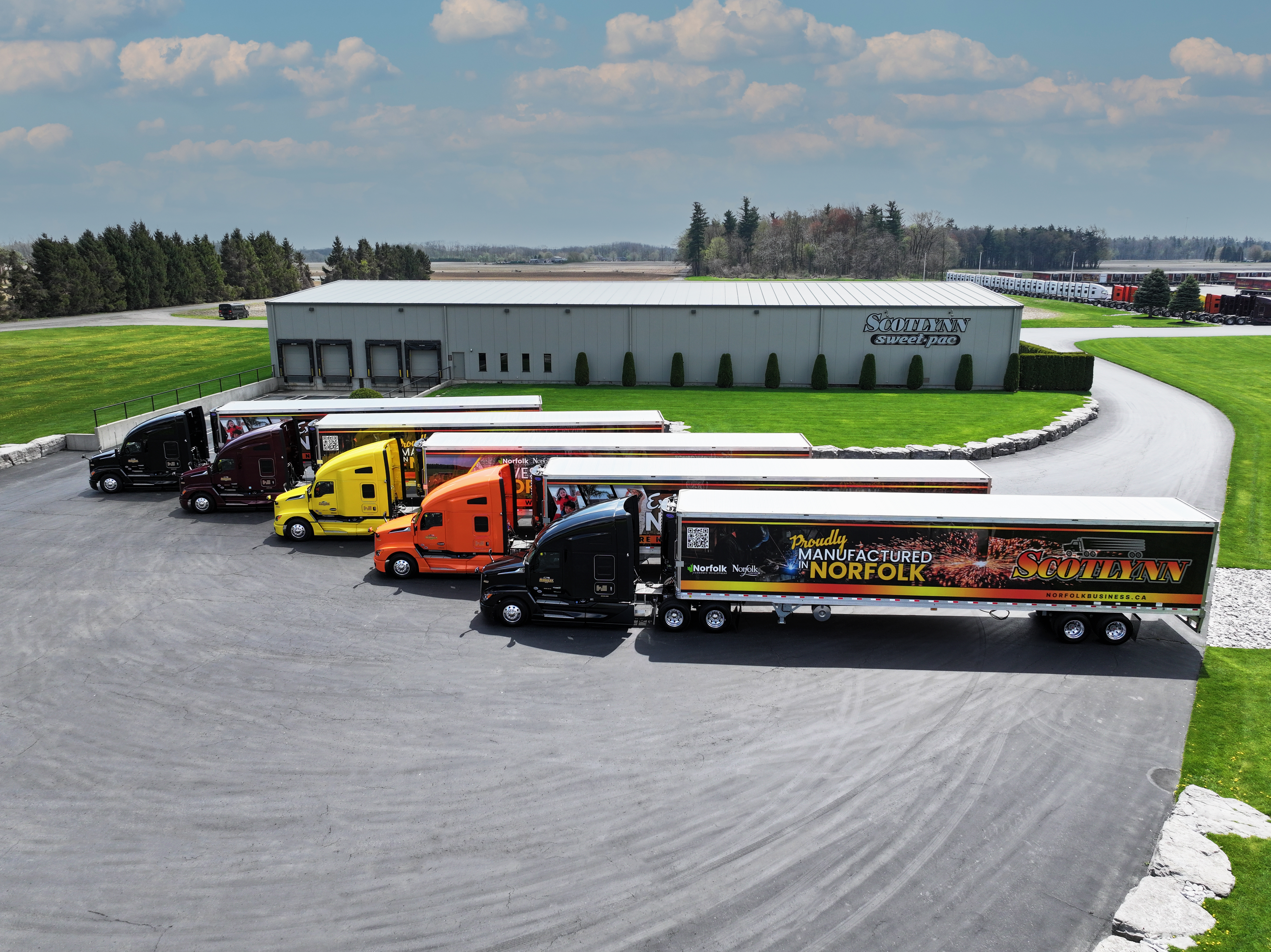Five Scotlynn trucks parked in front of the Scotlynn Sweetpac building, each displaying one of three Norfolk County promotional trailer designs.