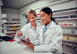 An image of two female pharmacists in white coats looking down at an iPad behind a pharmacy counter.