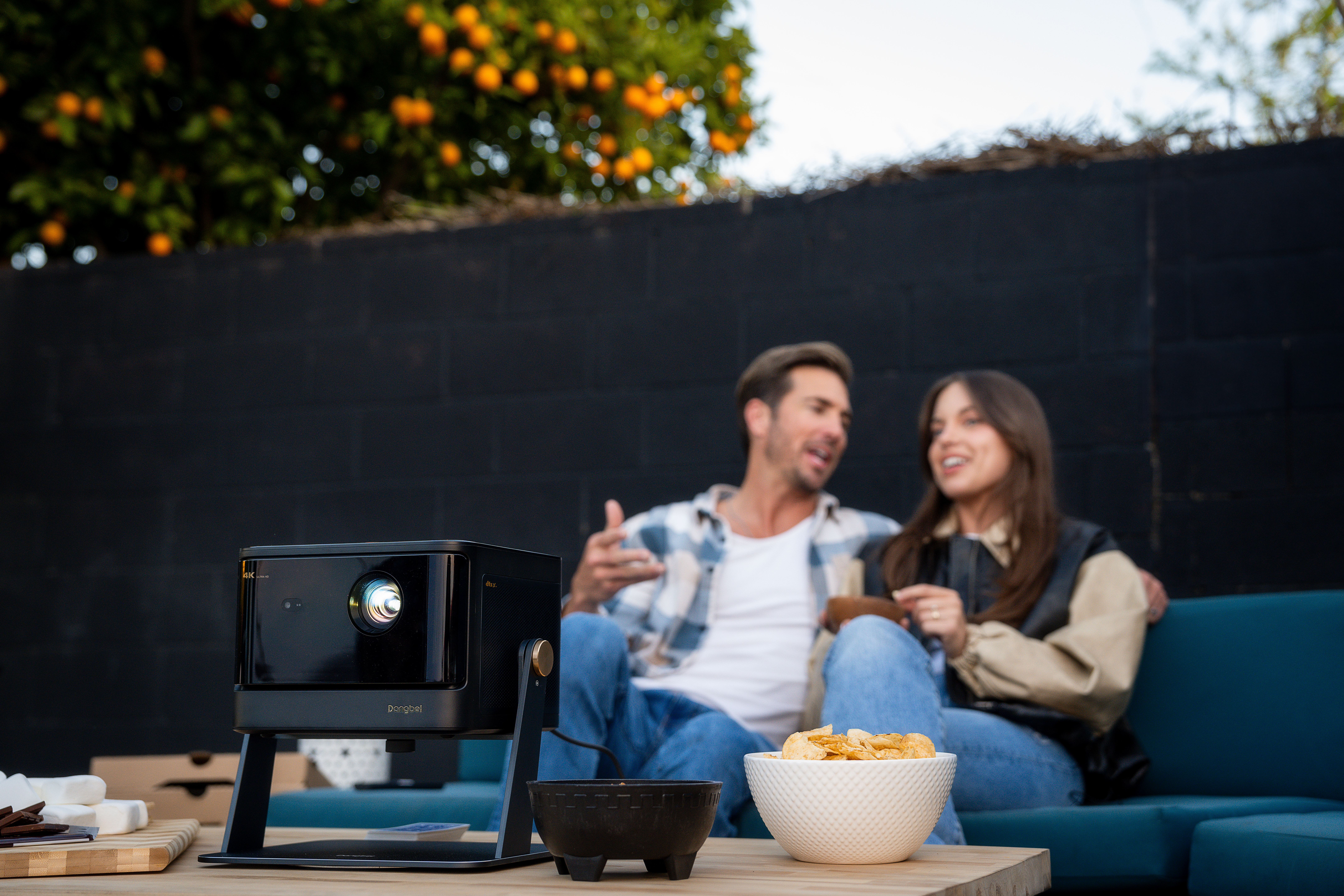 Dangbei DBOX02 projector on a table with snacks in front of a couple enjoying an outdoor movie night.