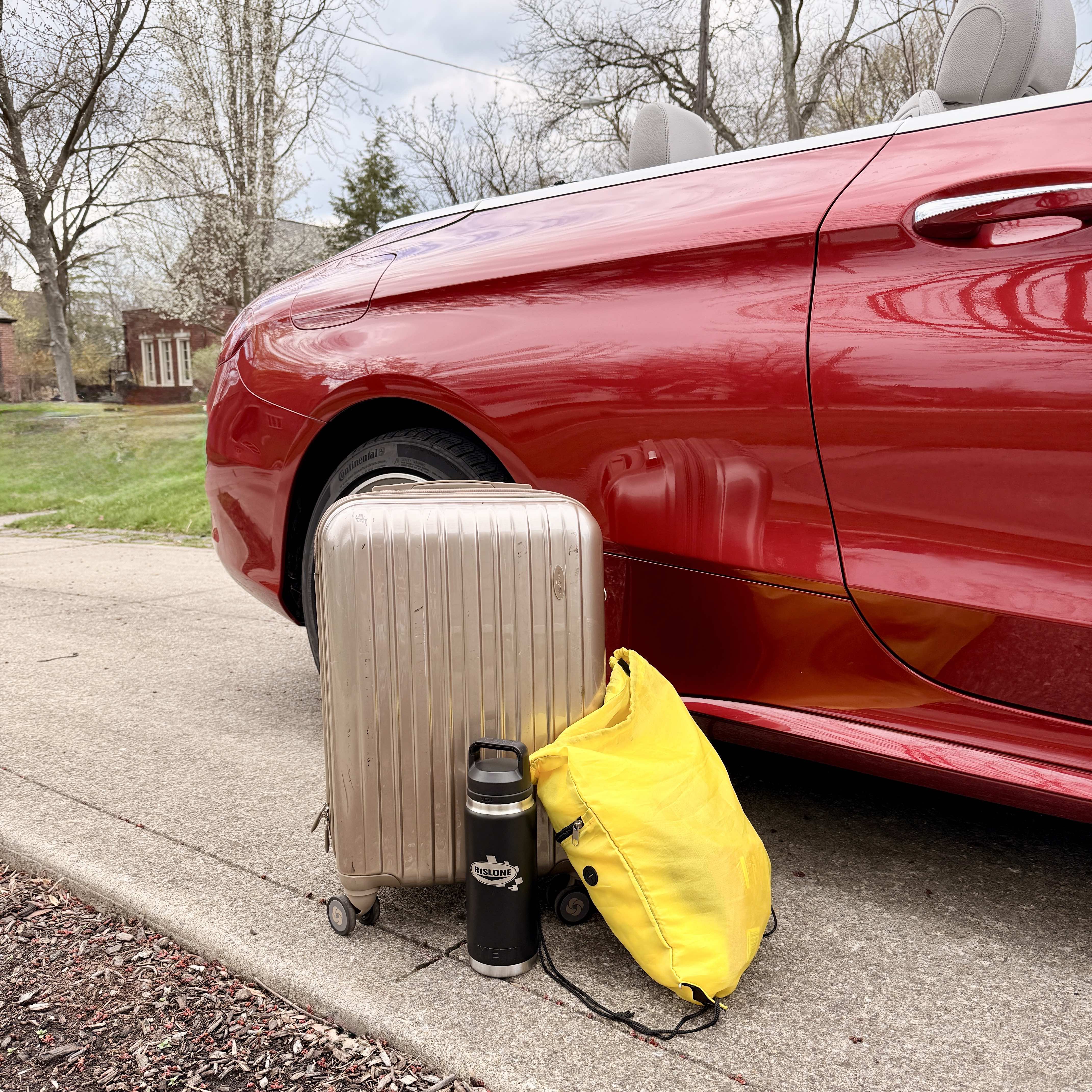 A suitcase, backpack and Rislone water bottle sit near the fuel door of a red convertible. 