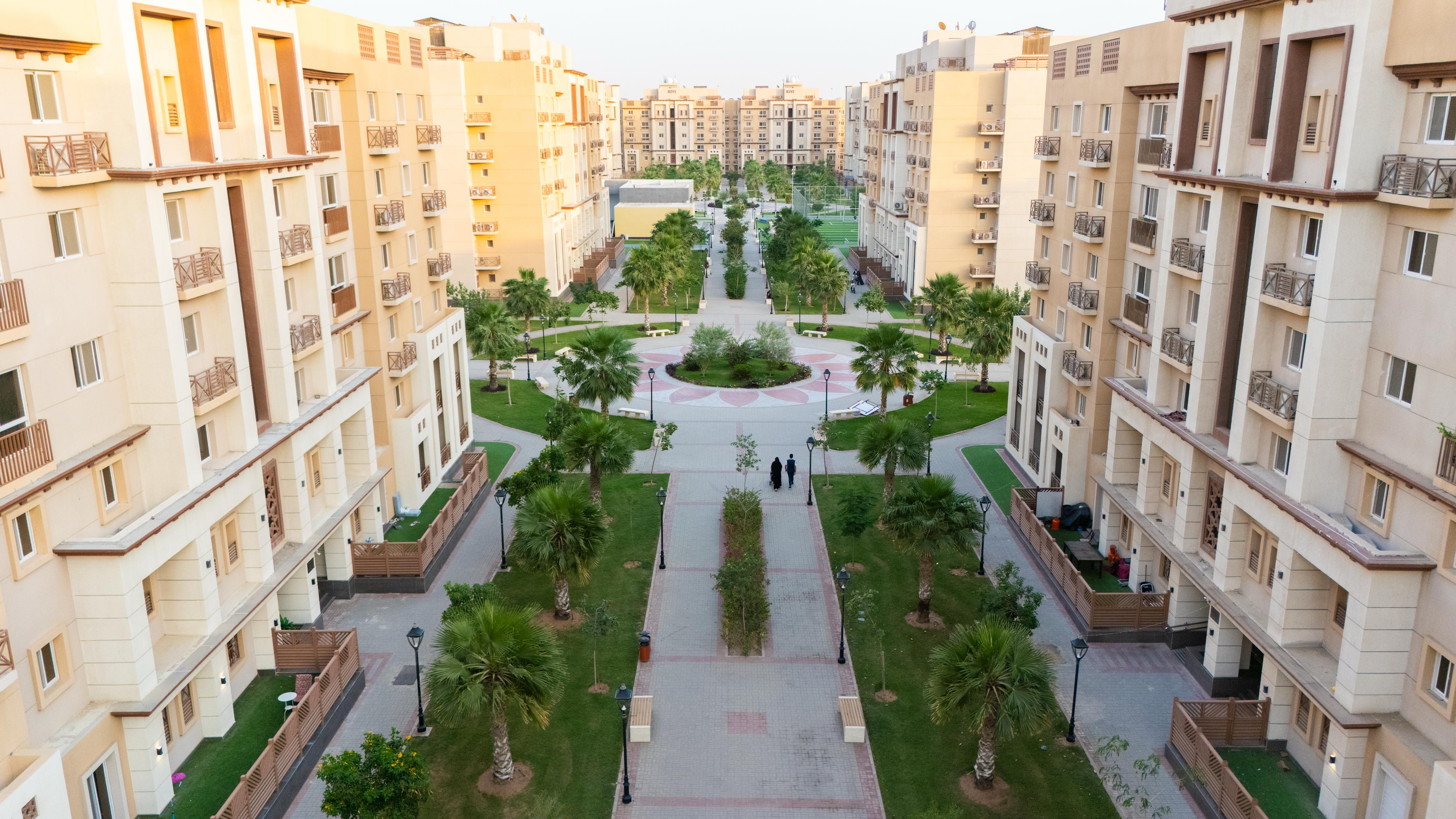 A wide aerial view of a residential complex with symmetrical buildings, central green walkways, palm trees, and people walking.