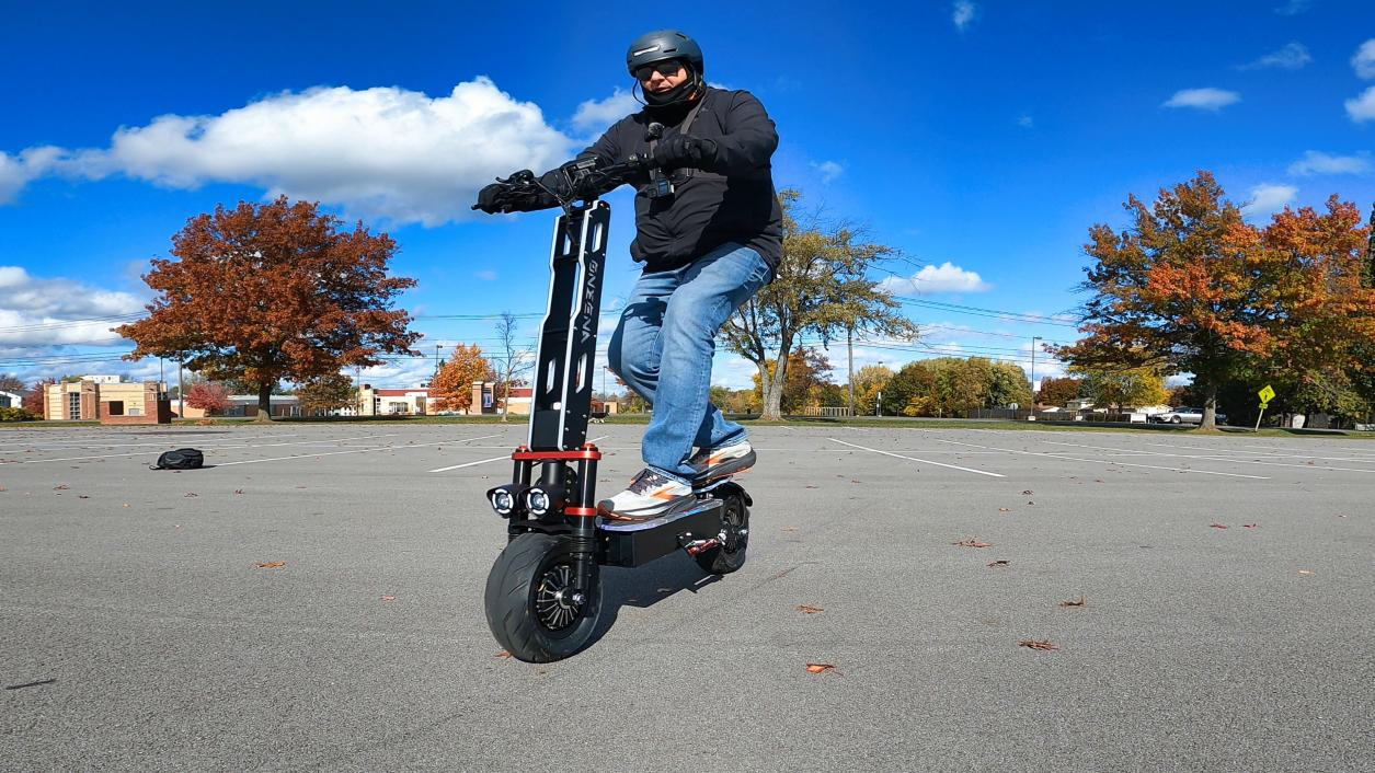 Helmeted rider on the ONECNA GT9 electric scooter, headlights on in a sunny parking lot.