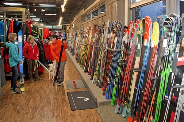 A long wall full of hundreds of pairs of skis and two people shopping for ski gear with a dog
