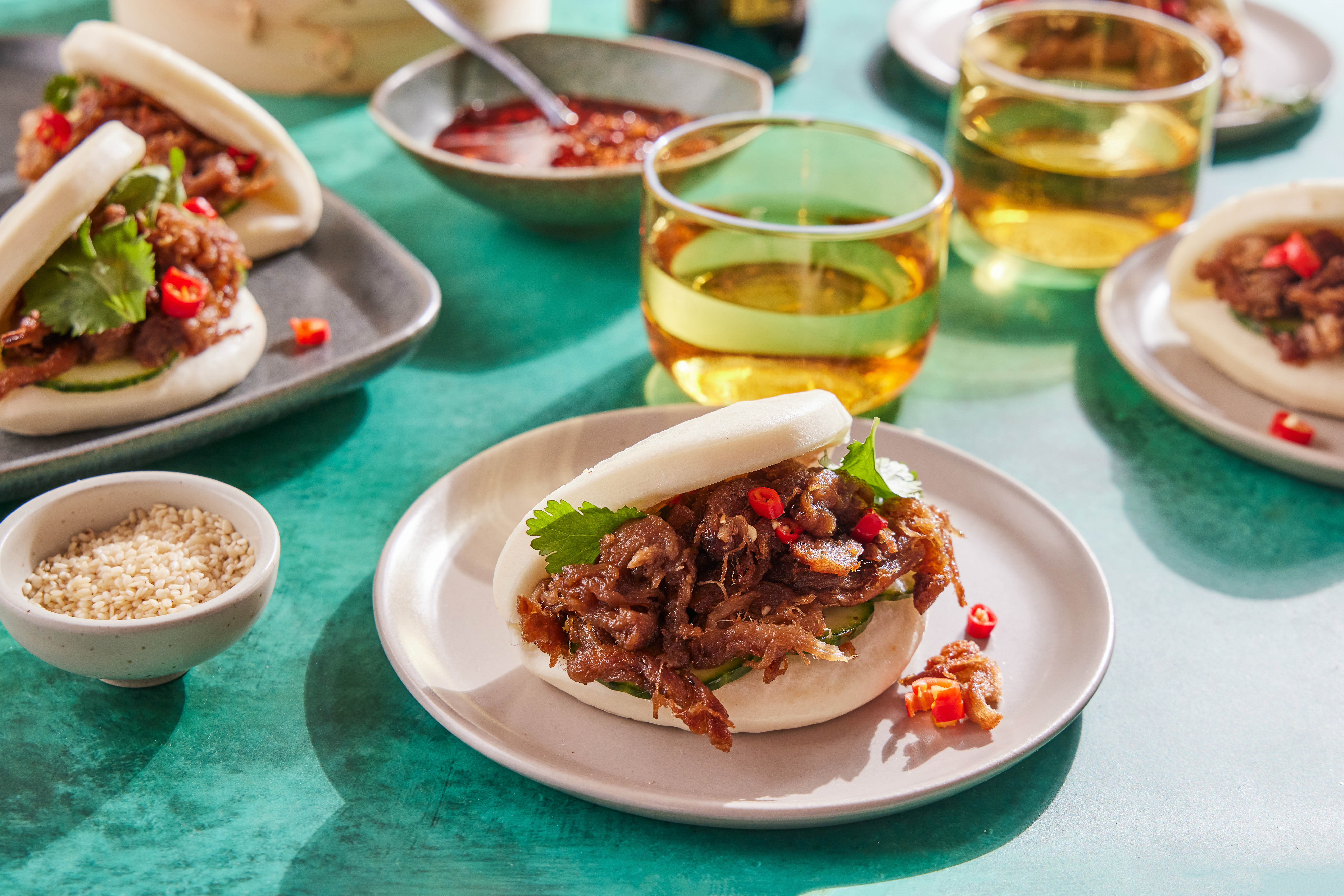 Tender™ plant-based shredded pork bao buns on a gray plate, accompanied by a bowl of chili sauce and glasses of tea on a teal table.