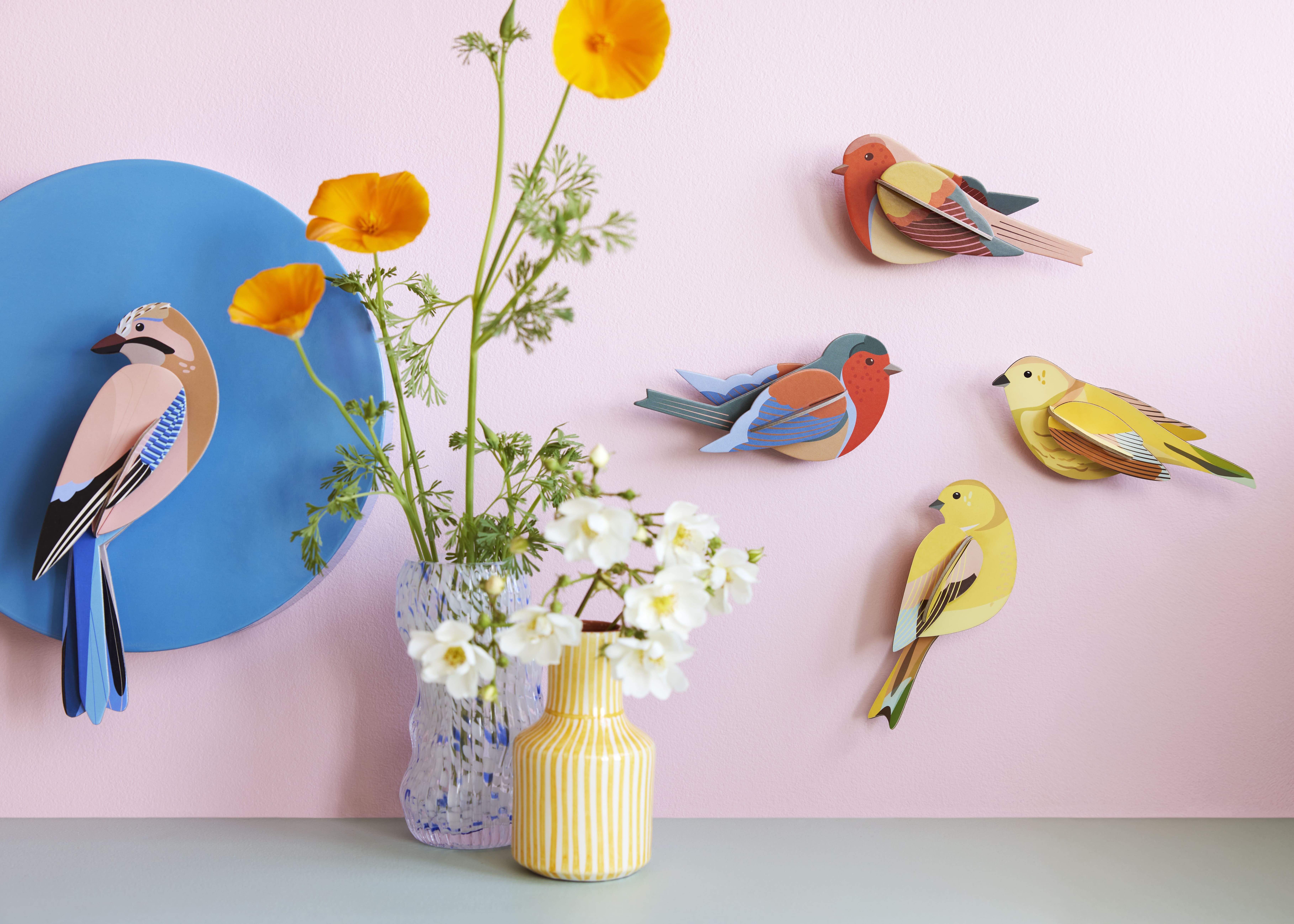 Oiseaux 3D en carton recycl de Studio ROOF accrochés sur un mur rose, accompagnés de vases fleuris.