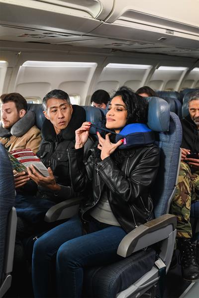 Images shows a woman on an airplane wearing a TNE S3 neck travel pillow from Cabeau with other passengers looking on in envy.