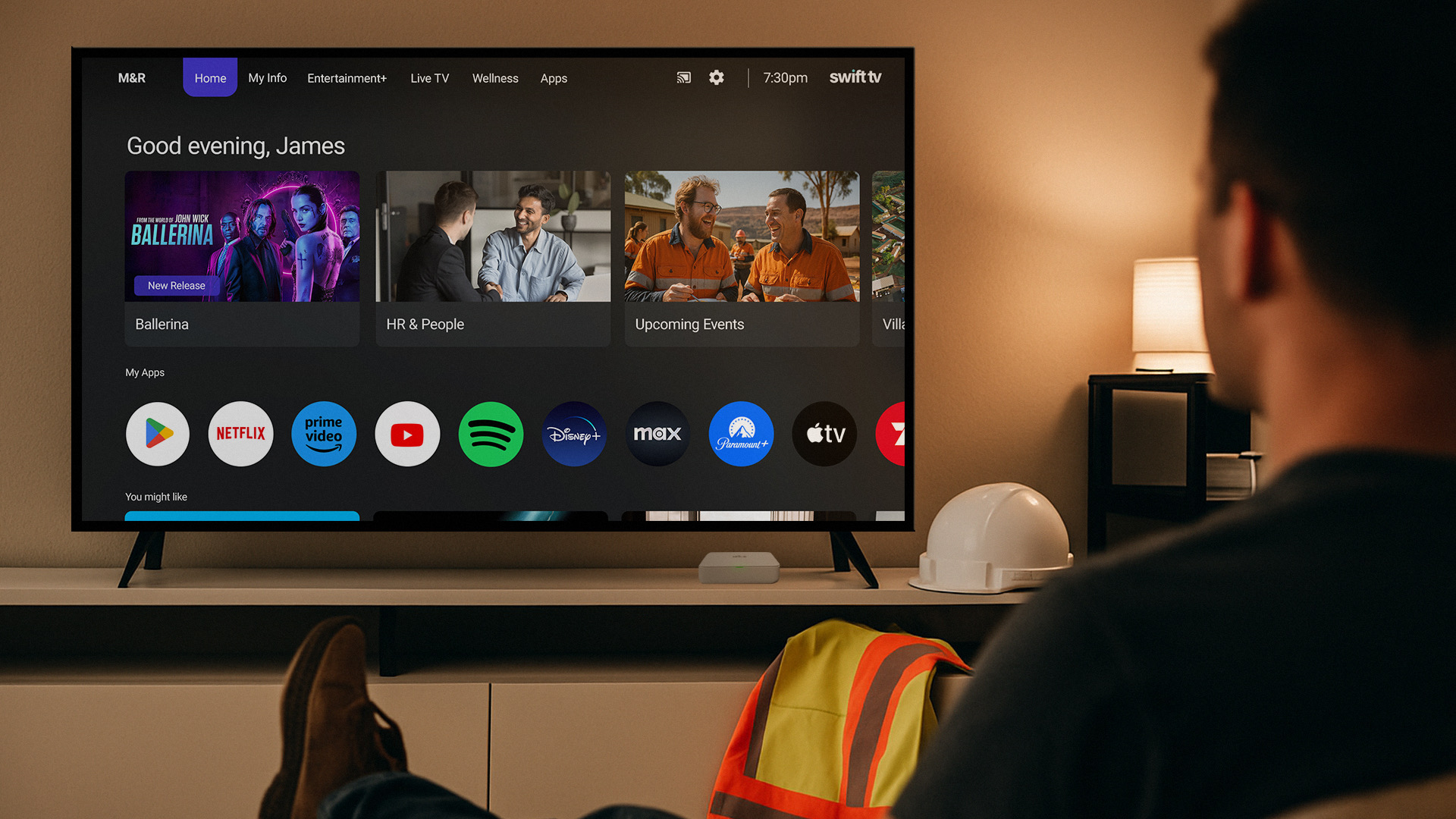 A man in work boots sits facing a TV screen showing the Swift TV interface, with apps like Netflix and Prime Video alongside company content.